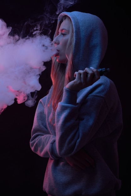 Retrato de hermosa chica rubia en sudadera con capucha gris fuma vape aislado sobre fondo negro de estudio, nube de humo de vapor, mini cachimba