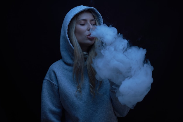 Retrato de hermosa chica rubia en sudadera con capucha gris fuma vape aislado sobre fondo negro de estudio, nube de humo de vapor, mini cachimba