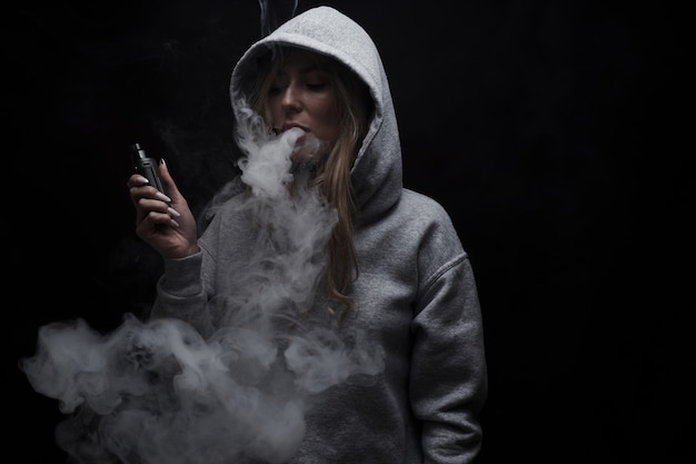 Retrato de hermosa chica rubia en sudadera con capucha gris fuma vape aislado sobre fondo negro de estudio, nube de humo de vapor, mini cachimba