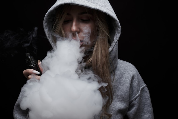 Retrato de hermosa chica rubia en sudadera con capucha gris fuma vape aislado sobre fondo negro de estudio, nube de humo de vapor, mini cachimba