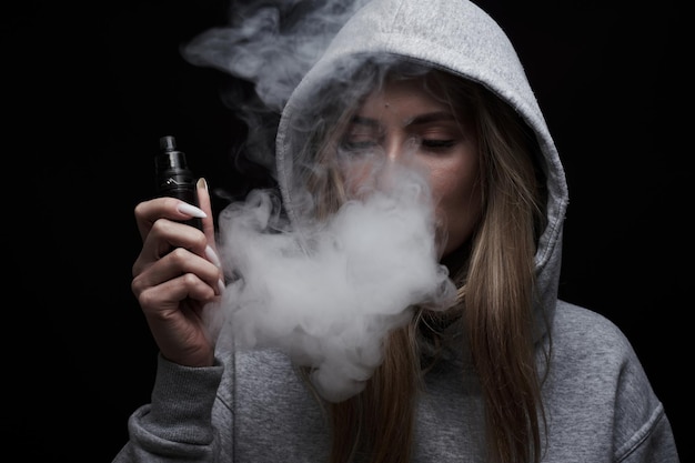 Retrato de hermosa chica rubia en sudadera con capucha gris fuma vape aislado sobre fondo negro de estudio, nube de humo de vapor, mini cachimba