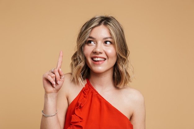 Retrato de una hermosa chica rubia con un vestido que se encuentran aisladas sobre la pared beige, señalar con el dedo