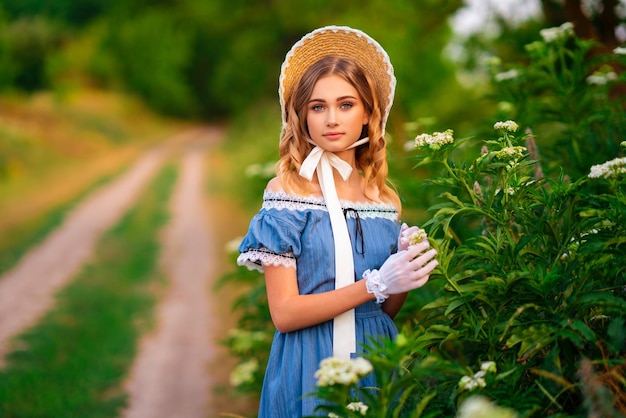 Retrato de una hermosa chica con un vestido vintage azul y un sombrero de paja cerca de las flores.
