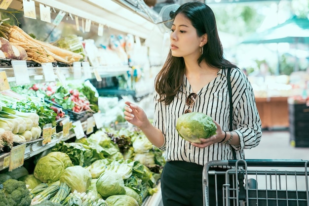 retrato hermosa dama urbana asiática está seleccionando coles frescas con concentración en la sección de productos mientras compra en el supermercado local.