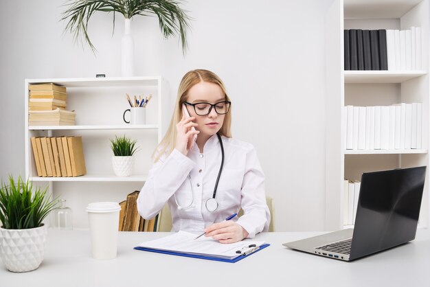 Retrato de una hermosa doctora sonriente mientras habla por el teléfono inteligente y completa una información médica