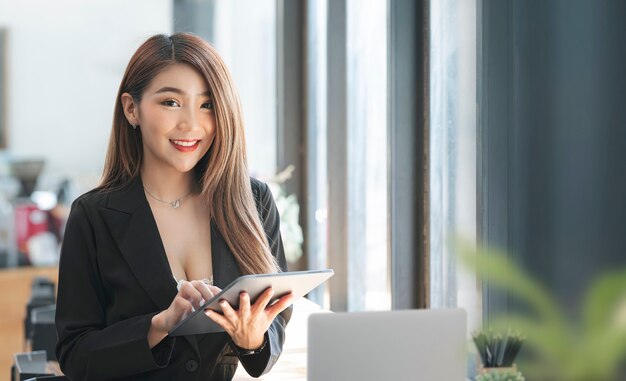 Foto retrato de hermosa empresaria sonriendo y mirando a cámara mientras trabaja con tableta en su oficina.