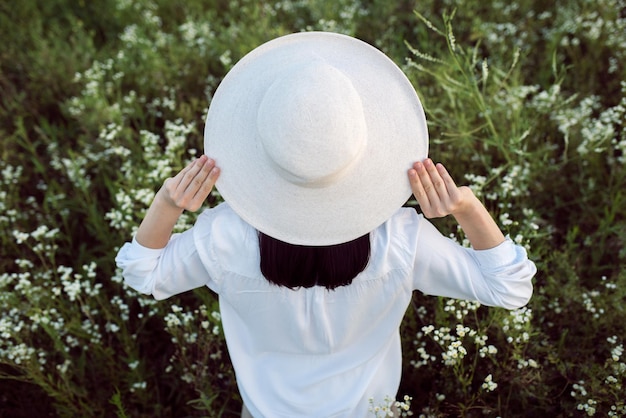 Retrato de hermosa y encantadora vista trasera de una joven morena bonita con las manos en un sombrero en el fondo del campo de flores