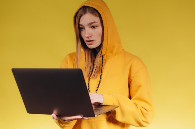 Retrato de una hermosa estudiante que trabaja con una computadora portátil La niña está vestida con una sudadera con capucha amarilla Foto sobre un fondo amarillo