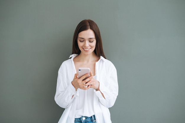 Retrato de una hermosa estudiante sonriente con cabello largo oscuro en camisa blanca usando un teléfono móvil en la mano sobre fondo gris generación aislada Z