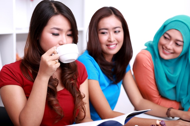 Retrato de hermosa estudiante universitaria bebiendo una taza de té y leyendo un libro mientras sus dos amigos la miran