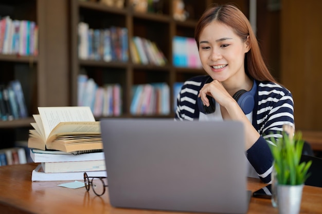 Retrato de una hermosa estudiante viendo y escuchando videos tutoriales en línea con auriculares y una computadora portátil sentada en una mesa en la biblioteca