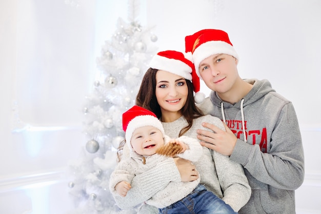 Retrato de hermosa familia alegre en gorros rojos y suéteres riendo y sonriendo al frente abrazando contra el árbol de Navidad decorado