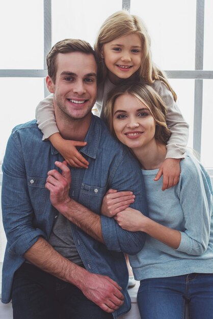 Foto retrato de una hermosa familia joven mirando a la cámara sonriendo y abrazándose mientras se sienta cerca de la ventana en casa