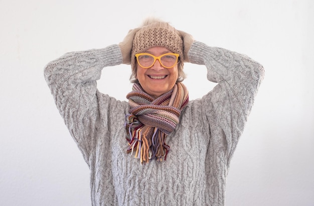 Retrato de una hermosa y feliz anciana con suéter gris usando anteojos y gorra mirando a la cámara aislada de fondo blanco