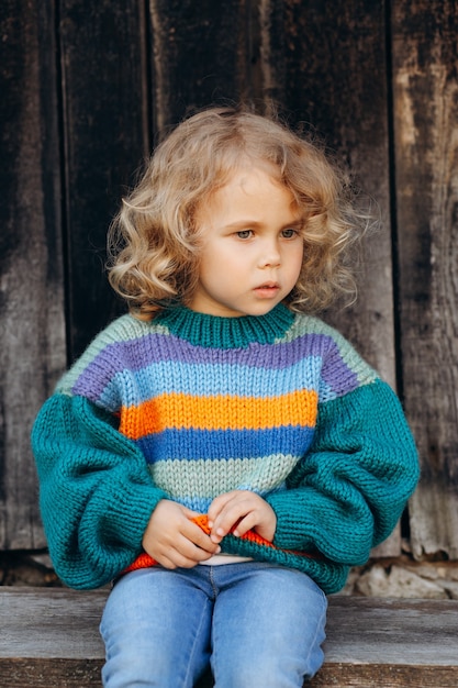 Retrato de una hermosa y feliz niña rizada en un suéter de punto cerca de una pared de madera