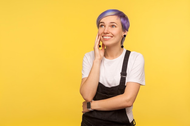 Retrato de una hermosa hipster feliz con el pelo corto violeta en overoles de mezclilla sosteniendo la palma cerca de la cara y soñando fantasías agradables, dulces recuerdos. aislado sobre fondo amarillo, foto de estudio