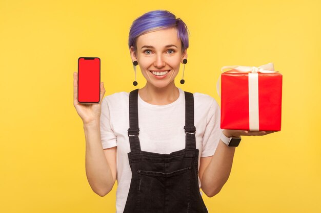 Retrato de una hermosa hipster sonriente con el pelo corto violeta en overoles sosteniendo una caja de regalo y un teléfono celular con pantalla roja simulada para publicidad aislada en una foto de estudio de fondo amarillo