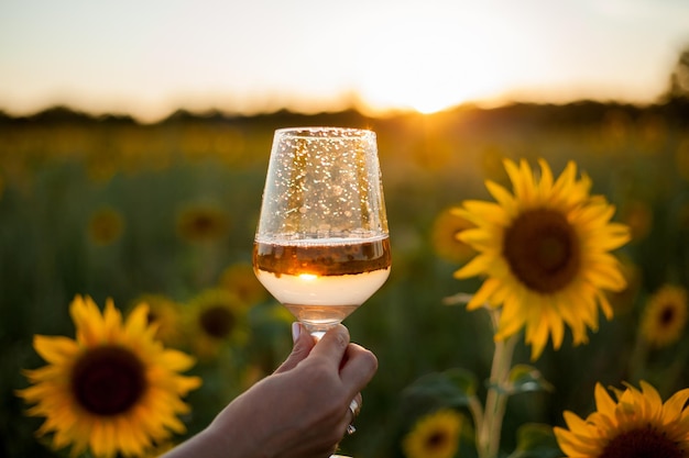 Retrato de una hermosa joven de 33 años con sombrero en el campo de girasoles al atardecer sostiene una copa de vino blanco en la mano Modelo feliz con vestido blanco en una noche de verano en la naturaleza Cálido