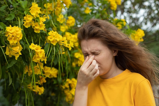 Retrato de una hermosa joven alérgica que sufre de alergia al polen o frío en el fondo de un árbol floreciente natural en primavera o en un día soleado de verano estornuda soplando su nariz mocosa frota los ojos