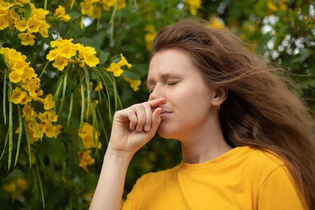 Retrato de una hermosa joven alérgica que sufre de alergia al polen o frío en el fondo de un árbol floreciente natural en primavera o en un día soleado de verano estornuda soplando su nariz mocosa frota los ojos