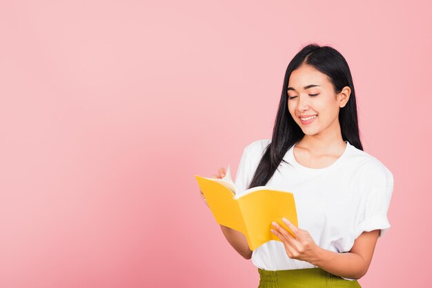Retrato de una hermosa joven asiática adolescente sonriendo sosteniendo un libro amarillo y leyendo, foto de estudio aislada en un fondo rosa con espacio para copiar