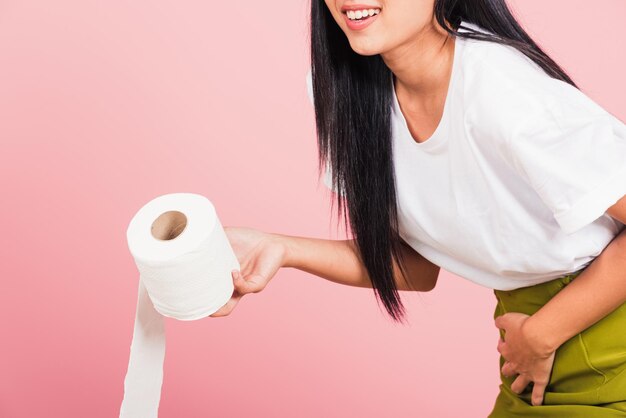 Foto retrato de una hermosa joven asiática diarrea estreñimiento sosteniendo dolor de estómago y rollo de papel higiénico en la mano ella entrepierna parte inferior del abdomen quiere ir al baño, tiro de estudio aislado en fondo rosa