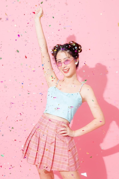 Retrato de una hermosa joven asiática feliz con un lindo vestido azul de moda que usa lentes de sol con las manos levantadas para celebrar la caída de la mosca de confeti en un espacio aislado de fondo de color rosa pastel espacio vacío Concepto de fiesta