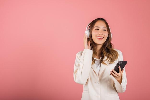 Retrato de una hermosa joven asiática que se divierte escuchando música con auriculares en una aplicación de teléfono inteligente, una mujer feliz que usa un teléfono móvil escucha una canción fuerte, una toma de estudio aislada en un fondo rosado