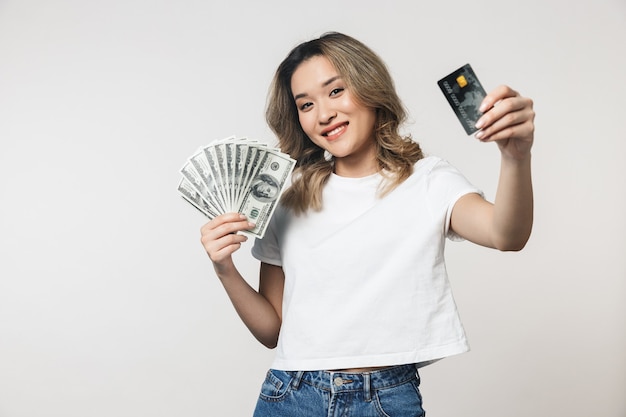 Retrato de una hermosa joven asiática que se encuentran aisladas sobre una pared blanca, mostrando dinero billetes y tarjetas de crédito de plástico