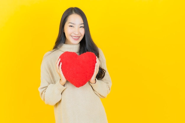 Retrato hermosa joven asiática sonríe con forma de almohada de corazón en la pared amarilla