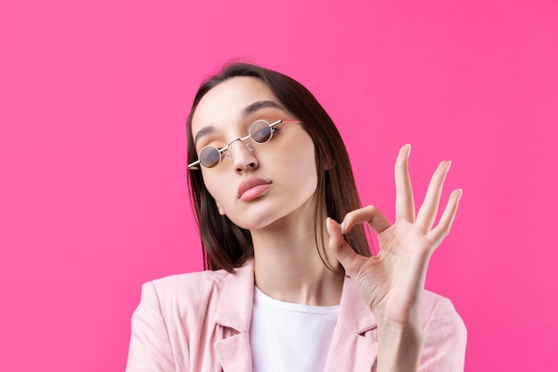 Retrato de una hermosa joven con una chaqueta rosa pensando aislada en un fondo rojo Muestra un signo OK