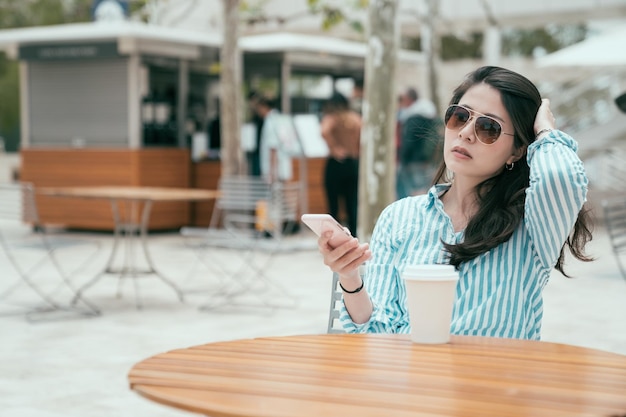 retrato de una hermosa joven china asiática leyendo un mensaje de texto en un teléfono móvil en una cafetería al aire libre. dama con gafas de sol disfruta del sol sentada en la mesa con una bebida para llevar en un vaso desechable