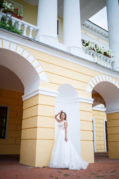 Retrato de una hermosa joven en el día de la boda de la novia, vestida con un vestido de novia blanco sobre un fondo de una gran casa con columnas