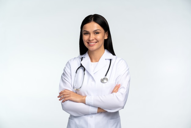 Foto retrato de hermosa joven doctora en chaqueta médica blanca aislada sobre fondo blanco. médico mujer morena cruzó las manos y sonriendo a la cámara