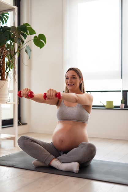 Retrato de una hermosa joven embarazada sentada y haciendo yoga.Usando pesas.