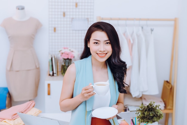 Retrato de hermosa joven empresaria asiática diseñadora de moda en su estudio mientras bebe café