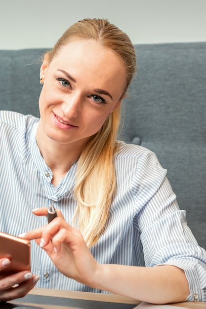 Retrato de hermosa joven empresaria caucásica con smartphone trabajando en oficina mirando y sonriendo a la cámara