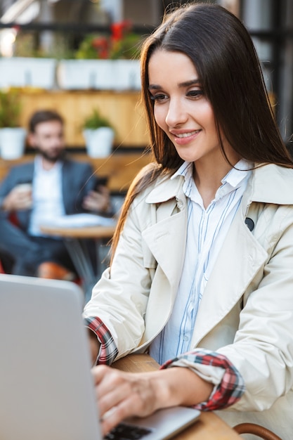 Foto retrato de hermosa joven empresaria en ropa formal sonriendo y trabajando en la computadora portátil mientras está sentado en el café al aire libre
