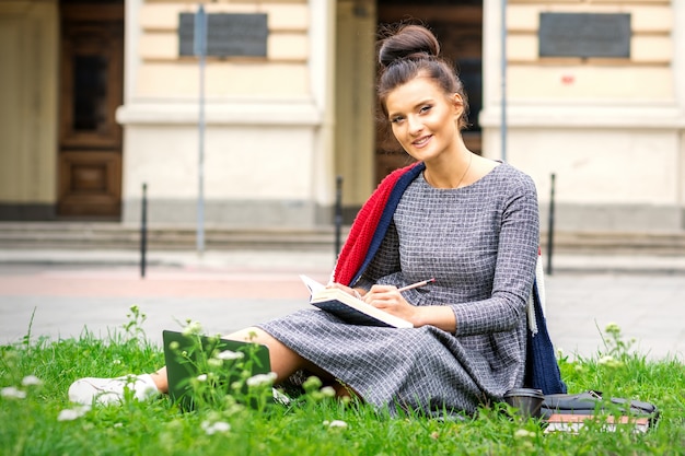 Retrato de una hermosa joven estudiante sonriente que estudia con un libro en el césped cerca de la universidad