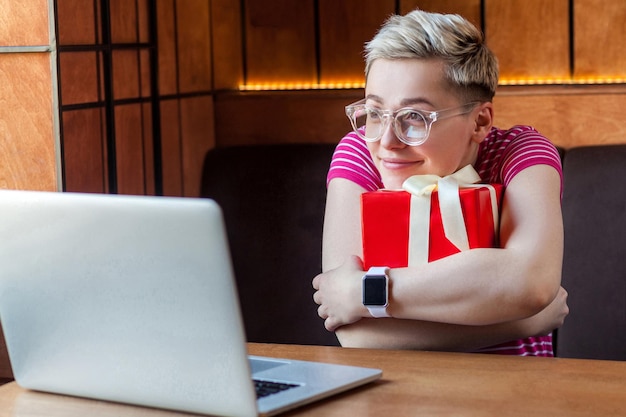 El retrato de una hermosa joven feliz soñando con el pelo corto en una camiseta rosa y anteojos está sentado en un café, abrazando una caja de regalo roja, mirando una laptop y sonriendo. Interior