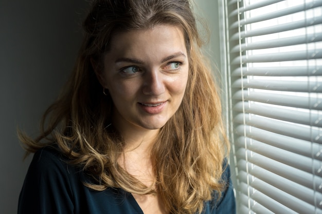 Retrato de una hermosa joven en una habitación sobre un fondo de una pared gris con sombras, luz natural desde la ventana.