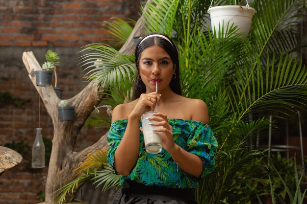 Retrato de una hermosa joven hispana con cabello largo y negro vestido casualmente bebiendo limonada de coco mirando a la cámara con una pared de ladrillos y plantas en el fondo Chica de belleza de estilo de vida