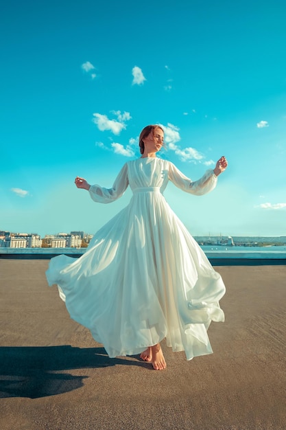 Retrato de una hermosa joven con un largo vestido blanco parada en la azotea de un edificio con paisaje urbano