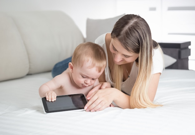 Retrato de hermosa joven madre acostada con su bebé en la cama y usando tableta