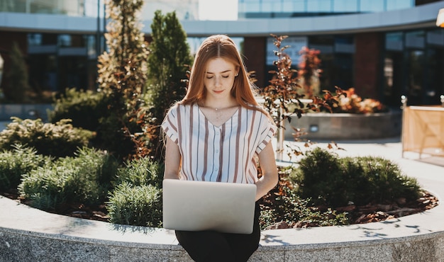 Retrato de una hermosa joven mirando su computadora portátil después de las lecciones. Joven estudiante haciendo los deberes en su computadora portátil al aire libre.