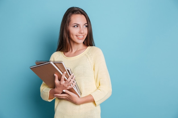 Retrato de hermosa joven morena sonriente sosteniendo cuadernos aislados en el fondo azul.