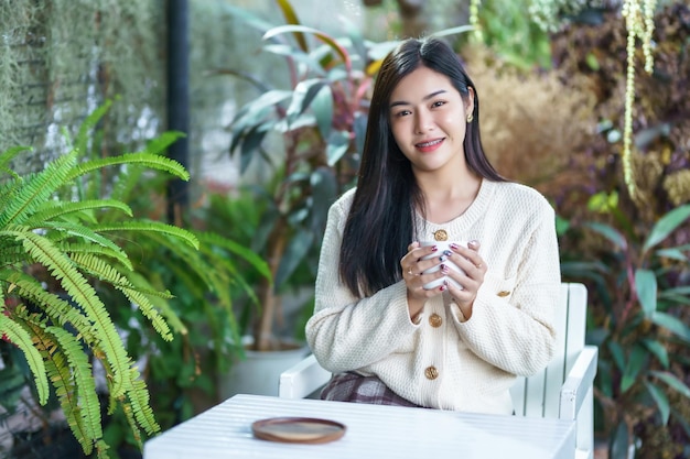 Retrato hermosa joven mujer asiática con ropa abrigada disfruta bebiendo sosteniendo de forma positiva la taza de café con leche o chocolate en casa en la sala de estar o en la cafetería