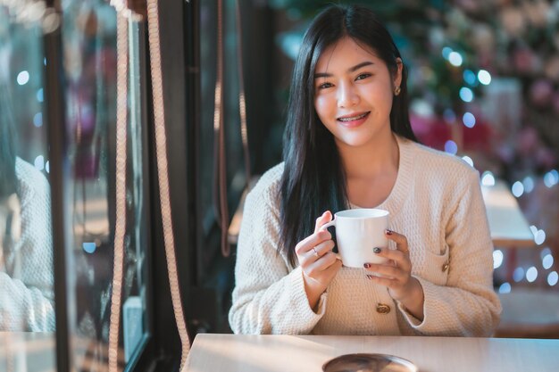 Retrato hermosa joven mujer asiática con ropa abrigada disfruta bebiendo sosteniendo positivamente la taza de café, leche o chocolate en casa en la sala de estar dentro o en la cafetería