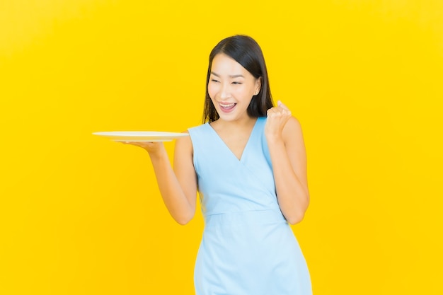 Retrato hermosa joven mujer asiática sonrisa con plato plato vacío en la pared de color amarillo