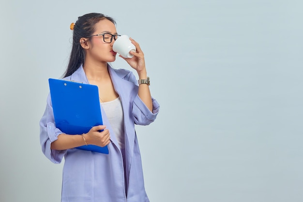 Retrato de una hermosa joven mujer de negocios asiática en traje disfrutando de una taza de café caliente y sosteniendo una carpeta de documentos aislada en un fondo morado